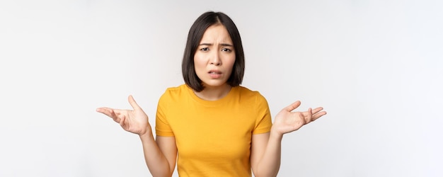 Une femme asiatique confuse et frustrée ne comprend pas qch qui a l'air désemparée en haussant les épaules debout en t-shirt jaune sur fond blanc