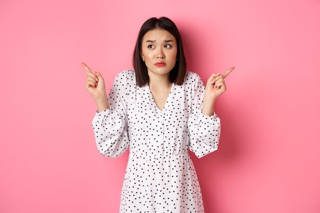 Femme asiatique confuse hésitant, haussant les épaules et pointant les mains des deux côtés, regardant indécise et ayant besoin d'aide pour choisir, debout sur fond rose