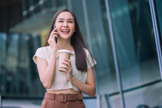 Une femme asiatique décontractée se détendre programmeuse nomade numérique regarde la conversation sur un smartphone tout en marchant dans la ville avec une tasse de café tenant un mode de vie souriant et joyeux sentiment positif rétrogradant