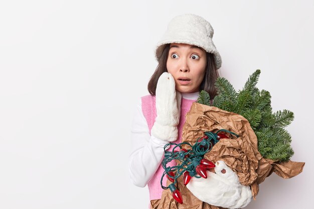 Une femme asiatique effrayée regarde impressionnée porte un pull panama et des mitaines porte des branches d'épinette à feuilles persistantes pour la décoration isolée sur fond blanc avec un espace de copie sur la gauche. Heure d'hiver.