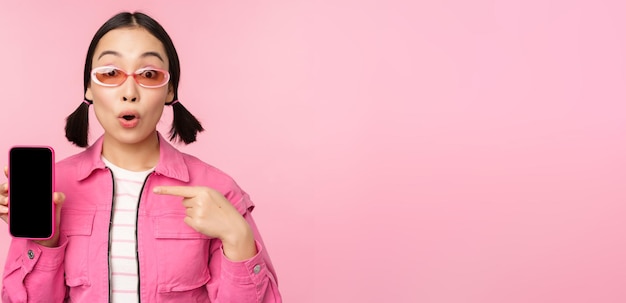 Femme asiatique enthousiaste dans des vêtements élégants lunettes de soleil pointant le doigt sur l'écran du téléphone mobile montrant l'application smartphone debout sur fond rose