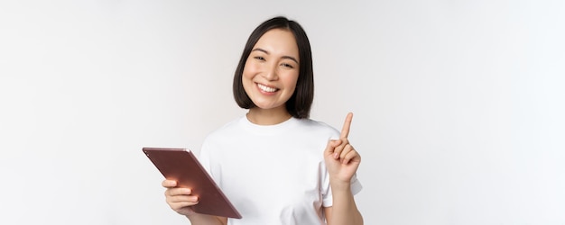 Femme asiatique enthousiaste avec tablette levant le doigt et regardant étonné pointant vers le haut debout sur whi