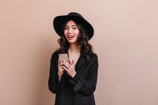 Femme asiatique étonnée tenant le smartphone et regardant la caméra. Élégante femme bouclée en manteau posant avec gadget.