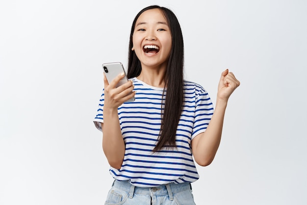 Femme asiatique excitée se réjouissant de la notification de l'application smartphone, gagnant sur téléphone mobile, debout sur blanc