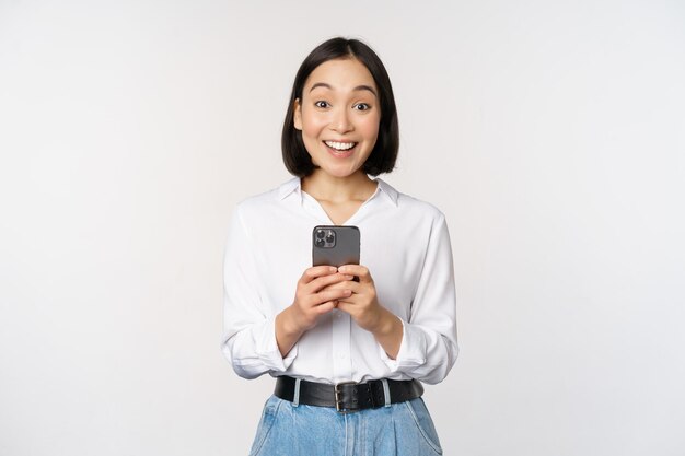 Femme asiatique excitée souriante réagissant aux informations sur le téléphone portable tenant le smartphone et regardant la caméra heureuse debout sur fond blanc