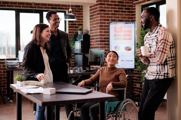 Femme asiatique en fauteuil roulant et groupe de personnes riant pendant la pause de travail, s'amusant dans un bureau adapté aux personnes handicapées. Travailleur souffrant d'un problème de santé chronique discutant avec des collègues joyeux.