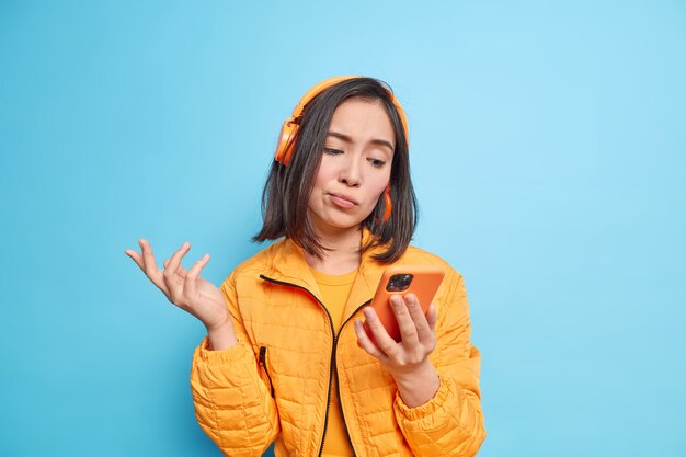 une femme asiatique hésitante perplexe regarde l'écran du smartphone ne peut pas choisir la chanson à écouter porte des écouteurs sans fil sur les oreilles porte une veste élégante isolée sur un mur bleu