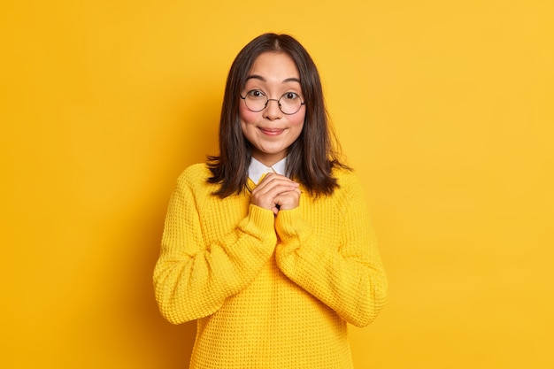 Une femme asiatique heureuse garde les mains jointes et anticipe quelque chose de bons sourires doucement a des fossettes sur les joues vêtues de vêtements décontractés.