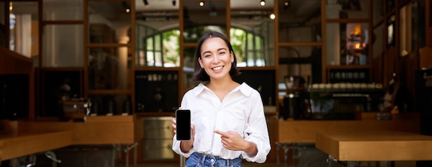 Photo gratuite une femme asiatique heureuse, gérante d'un café, se tient devant un restaurant et pointe du doigt une application pour smartphone.