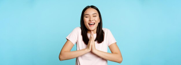 Une femme asiatique heureuse et souriante montre un geste de namaste mendiant et rêvant de quelque chose qui fait de grands espoirs