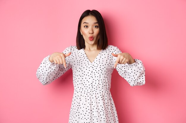 Femme asiatique impressionnée en robe pointant les doigts vers le bas, regardez là un geste, consultez les remises et les ventes, debout étonnée sur fond rose