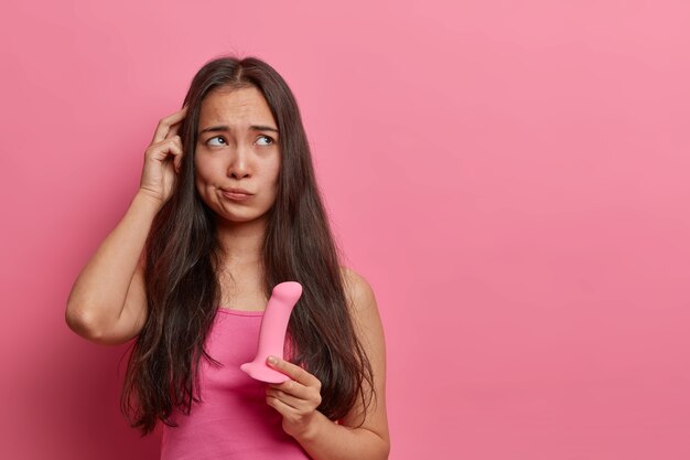 Une femme asiatique indécise se frotte la tête choisit un gode dans un sex-shop détient un objet phallique rose en silicone pour fournir un plaisir sexuel via la pénétration trouve la taille parfaite pour elle-même, remplace les organes génitaux de vrais hommes
