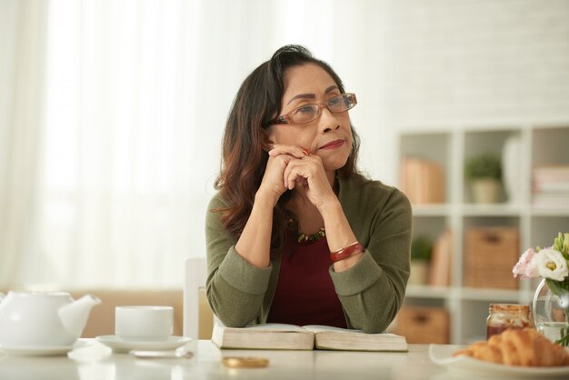 Femme asiatique mature pense à quelque chose assis à la table à la recherche de suite