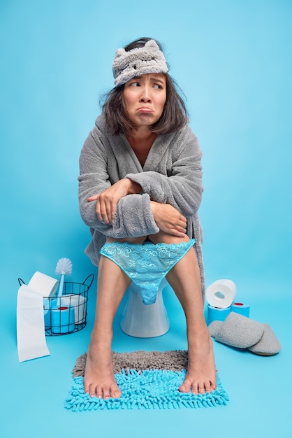Photo gratuite une femme asiatique mécontente se sent malheureuse et endormie après un réveil précoce porte un masque de sommeil et un pantalon en dentelle abaissé sur les jambes pose dans les toilettes sur le mur bleu de la cuvette des toilettes souffre de diarrhée