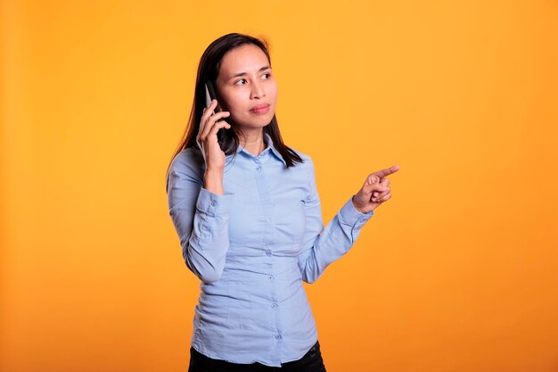 Femme asiatique parlant au téléphone portable avec un ami à distance, appréciant la conversation en studio sur fond jaune. Jeune adulte insouciant positif répondant à l'appel du smartphone. Notion de communication