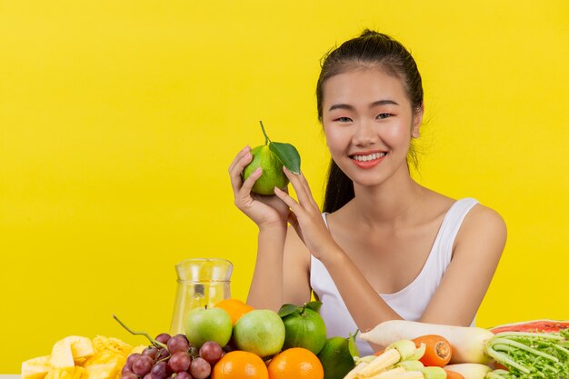 Une femme asiatique portant un débardeur blanc. Holdingorange avec la main droite Et sur la table il y a beaucoup de fruits différents.