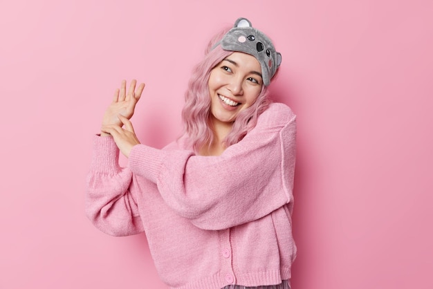 Une femme asiatique positive et insouciante aux cheveux teints danse et garde les sourires levés à la main largement a une expression rêveuse porte un pull décontracté masque de sommeil isolé sur fond rose. Concept d'émotions heureuses