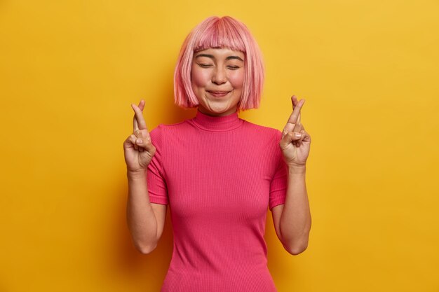 Une femme asiatique positive se croise les doigts avant un événement important sourit joyeusement, les yeux fermés, a de grands espoirs de mieux