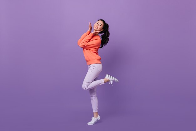 Femme asiatique raffinée debout sur une jambe. Vue sur toute la longueur de la femme chinoise heureuse dansant sur fond violet.