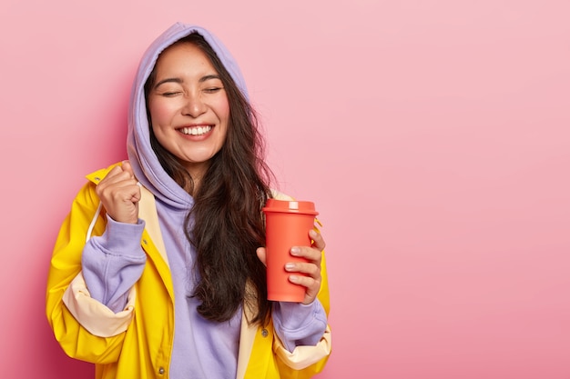 Une femme asiatique ravie de joie lève le poing fermé, garde les yeux fermés, boit une boisson chaude du flacon, se réjouit du moment heureux, porte des vêtements chauds et imperméables