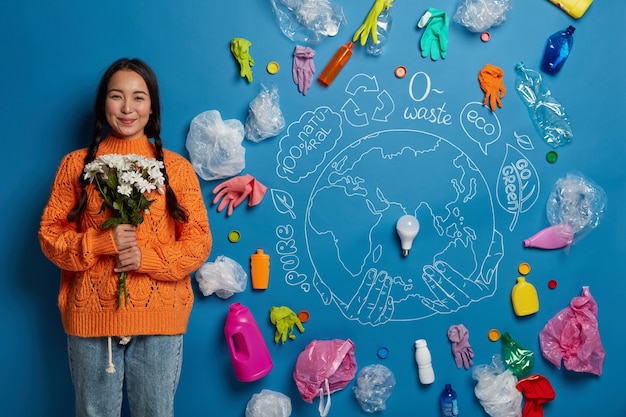 Femme asiatique satisfaite avec deux nattes, porte un pull orange, tient le bouquet, prend soin de la nature, est respectueuse de l'environnement, prête à recycler les déchets compilés.