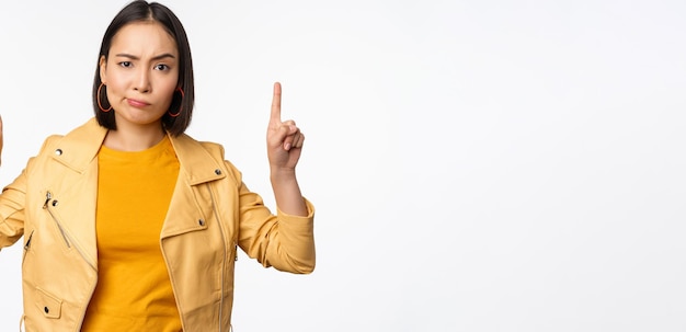 Femme asiatique sceptique pointant les doigts vers le haut en fronçant les sourcils et regardant avec désapprobation se plaindre de qch debout douteux sur fond blanc