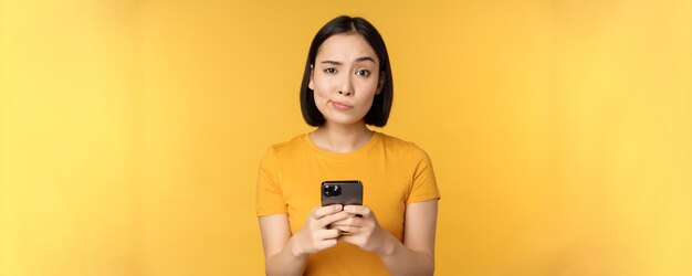 Femme asiatique sceptique tenant un smartphone regardant avec doute la caméra debout sur fond jaune