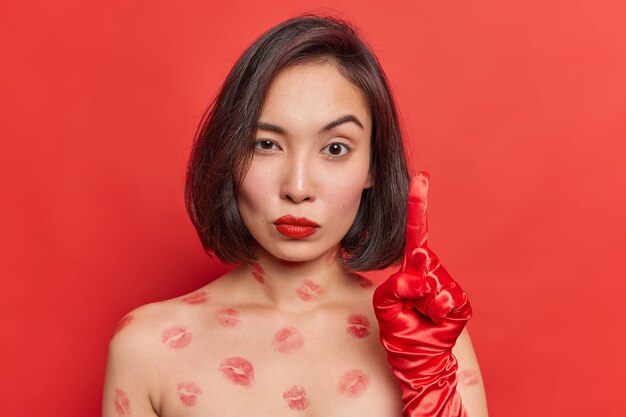 Une femme asiatique sérieuse et attentive lève l'index porte des gants se tient torse nu avec des traces de lèvres porte du rouge à lèvres rouge pose à l'intérieur a une excellente idée