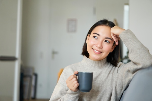 Photo gratuite femme asiatique souriante assise sur un canapé avec sa tasse buvant du café à la maison et se relaxant après le travail à la recherche de calme et de confort