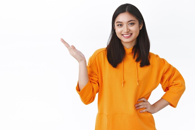 Femme asiatique souriante charismatique en sweat à capuche orange pointant sur la publicité, présente le produit avec une expression heureuse