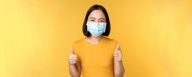 Femme asiatique souriante dans un masque médical montrant le pouce vers le haut comme une approbation et recommande qch debout sur fond jaune