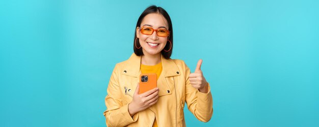 Femme asiatique souriante montrant le pouce levé enregistrant sur un téléphone mobile à l'aide d'une application pour smartphone et la recommandant debout sur fond bleu