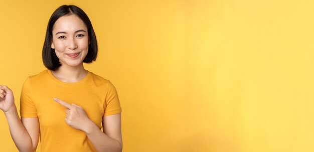 Femme asiatique souriante pointant les doigts vers la gauche montrant une publicité sur un espace de copie vide debout au-dessus de vous