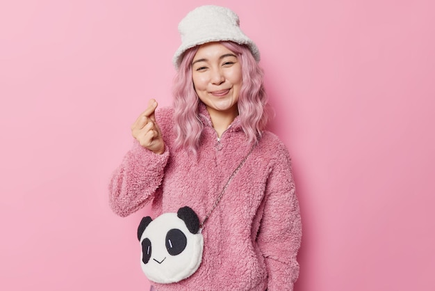 Une femme asiatique souriante et tendre aux cheveux roses montre un signe coréen comme des formes d'amour, un mini-cœur porte un manteau de fourrure et le panama a un panda comme un sac isolé sur fond rose. Notion de langage corporel