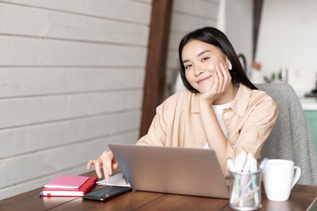 Femme asiatique souriante travaillant à distance à la maison, assise avec un ordinateur portable et un téléphone portable, une fille coréenne doin ...