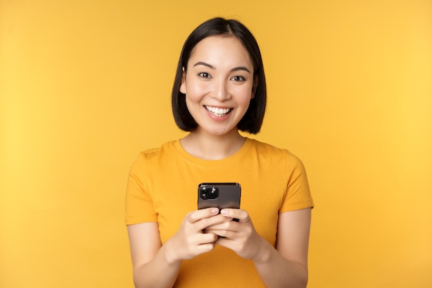 Femme asiatique souriante utilisant un téléphone portable tenant un smartphone dans les mains debout en t-shirt sur fond jaune