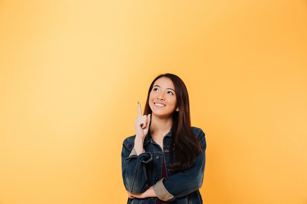 Femme asiatique souriante en veste en jean pointant et levant sur fond jaune