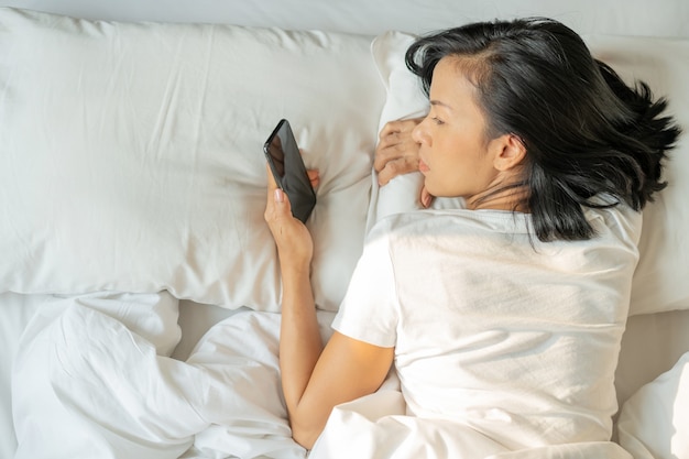 Femme asiatique avec un sourire attrayant utilise le smartphone tablette sur lit blanc. Vue de dessus