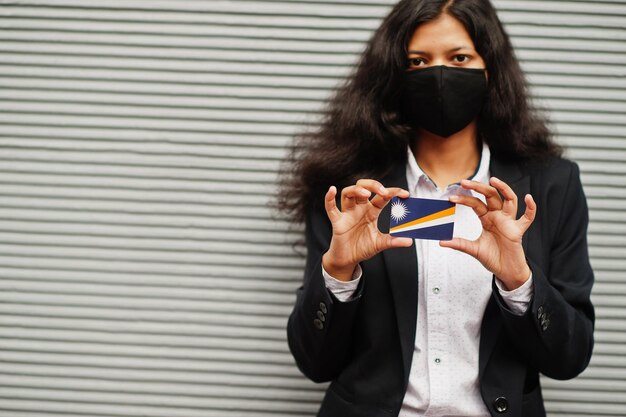 Une femme asiatique en tenue de soirée et un masque de protection noir tiennent le drapeau des Îles Marshall à portée de main sur fond gris Coronavirus au concept de pays