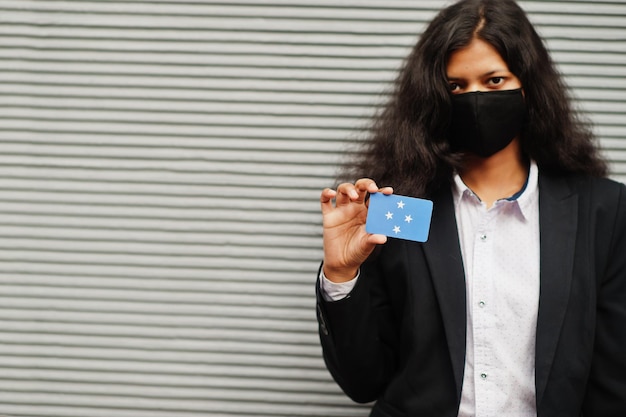 Une femme asiatique en tenue de soirée et un masque de protection noir tiennent le drapeau de la Micronésie à portée de main sur fond gris Coronavirus au concept de pays