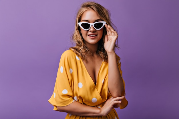 Femme assez bouclée dans des lunettes de soleil sombres posant. Modèle féminin sensuel en chemisier jaune debout sur violet.