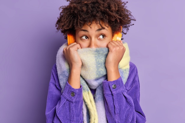 Photo gratuite une femme assez réfléchie se sent très froide après avoir marché par temps glacial porte un foulard couvre la moitié du visage tremble pendant une promenade en plein air porte un casque stéréo écoute de la musique habillée en chemise violette