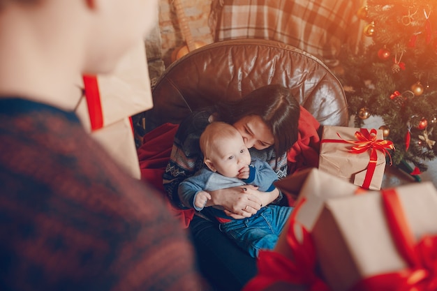 Femme Assise Sur Un Canapé Avec Un Bébé Dans Ses Bras Et Avec Une Montagne De Cadeaux Marron Avec Noeud Rouge