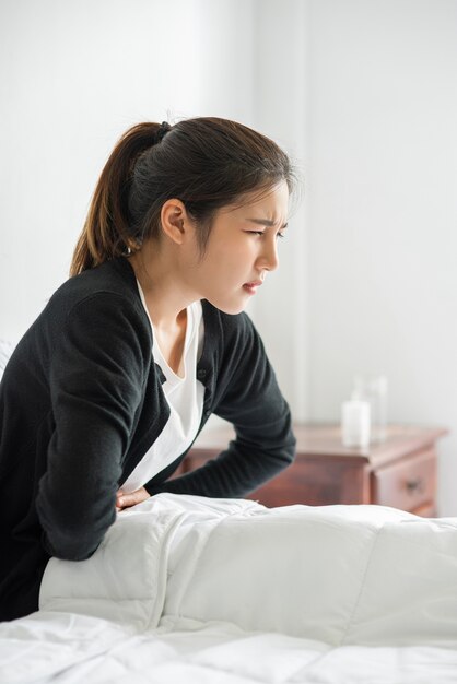 Une femme assise dans son lit avec des douleurs abdominales et en appuyant sa main sur son ventre.