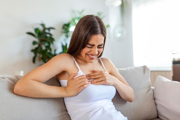 Femme assise avec une forte douleur à la poitrine et les mains touchant sa poitrine tout en ayant des problèmes à la maison Crise cardiaque ou symptôme d'insuffisance cardiaque