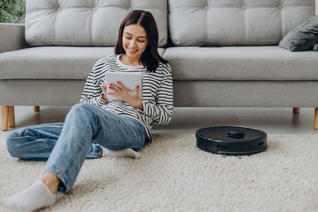 Femme assise près du canapé à l'aide d'une tablette pendant qu'un aspirateur robot nettoie la pièce