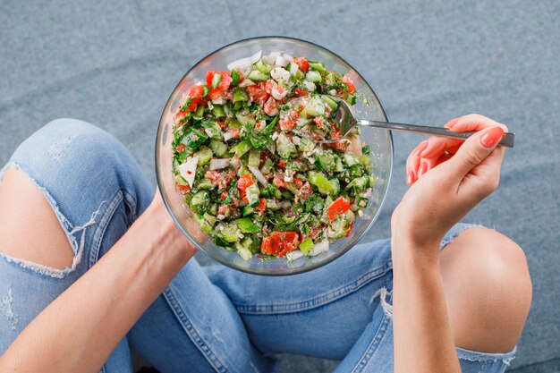 Femme assise sur le sol, tenant et mangeant une salade de légumes dans un bol en verre sur une vue de dessus de surface grise.