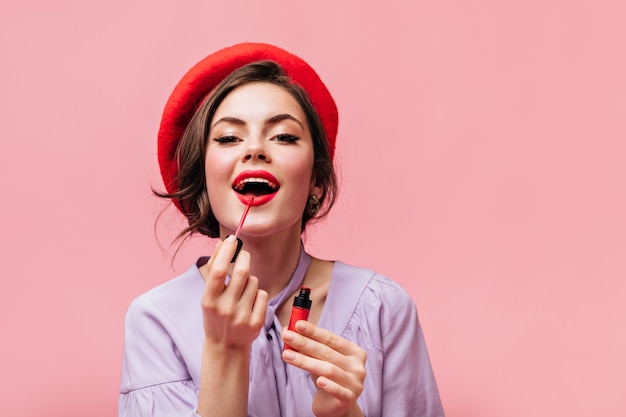 Femme au béret brillant peint ses lèvres avec du rouge à lèvres. Fille en chemisier lilas posant sur fond rose.