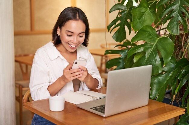 Femme au café assise avec un ordinateur portable à la surprise de l'écran du smartphone lisant de bonnes nouvelles passionnantes