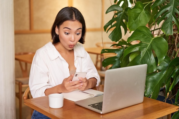 Femme au café assise avec un ordinateur portable à la surprise de l'écran du smartphone lisant de bonnes nouvelles passionnantes